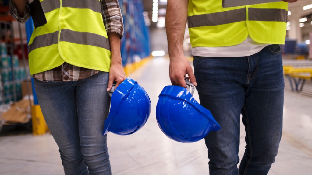 Deux travailleurs portant des gilets de haute visibilité et tenant des casques de protection bleus dans un entrepôt.